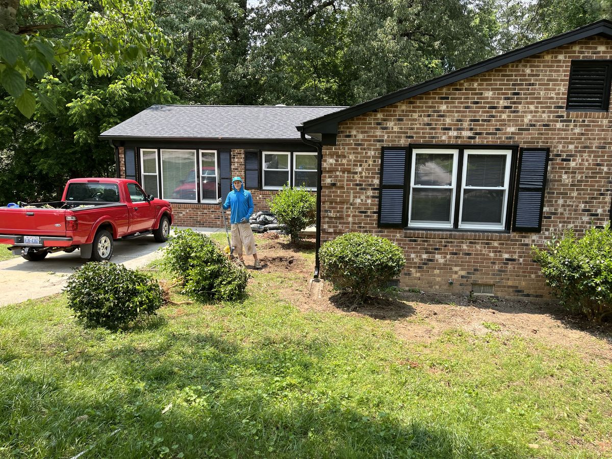 Addison in a blue sun hoodie with a shovel, planting shrubs along the front of the brick house